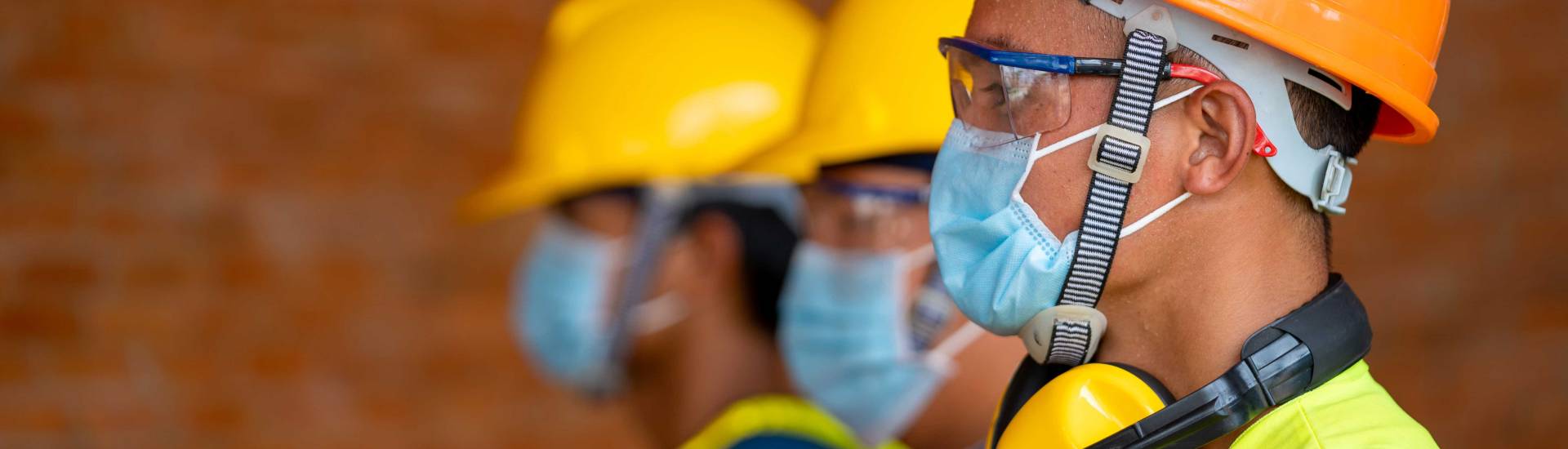 Three workers wearing safety helmet, goggles and face masks stand in a row.
